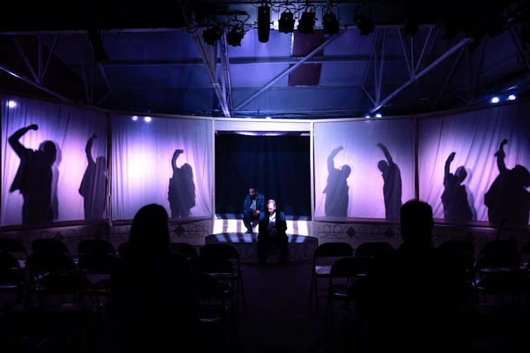 Photograph from the performance of Punishment Acts. It featuring two actors seated centre stage, to their left and right the shadows of actors with raised-arms cast shadows on illuminated white curtains. Dark silhouettes of seated audience members are visible in foreground, with stage lighting creating dramatic contrast and highlighting key figures.