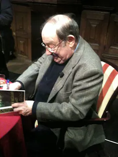 Un hombre con americana firma un libro sentado frente a una mesa.