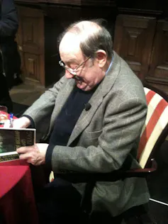 Un hombre con americana firma un libro sentado frente a una mesa.