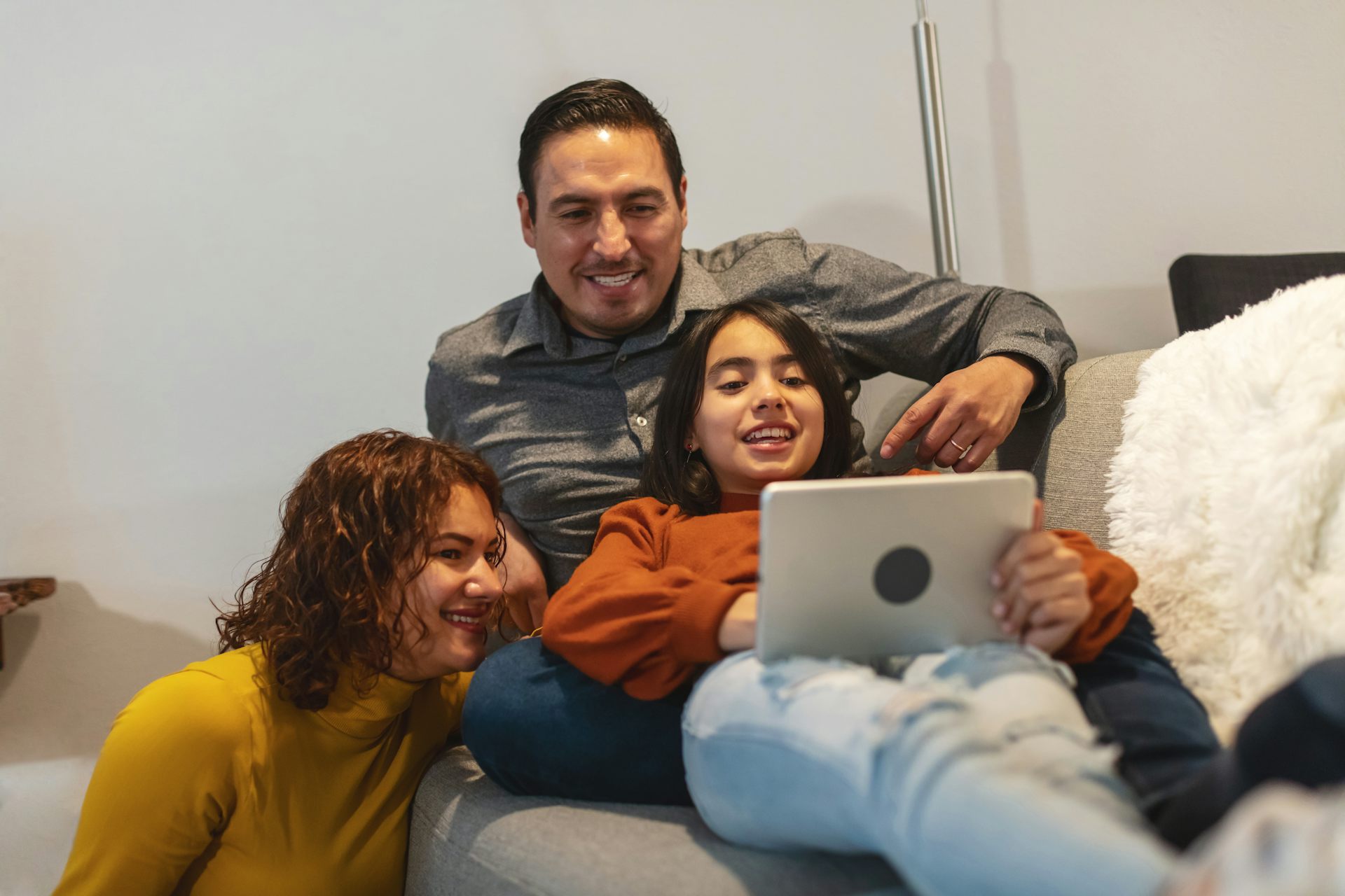 A middle-aged mom and dad sit close to their daughter, looking at the laptop on her lap and laughing.