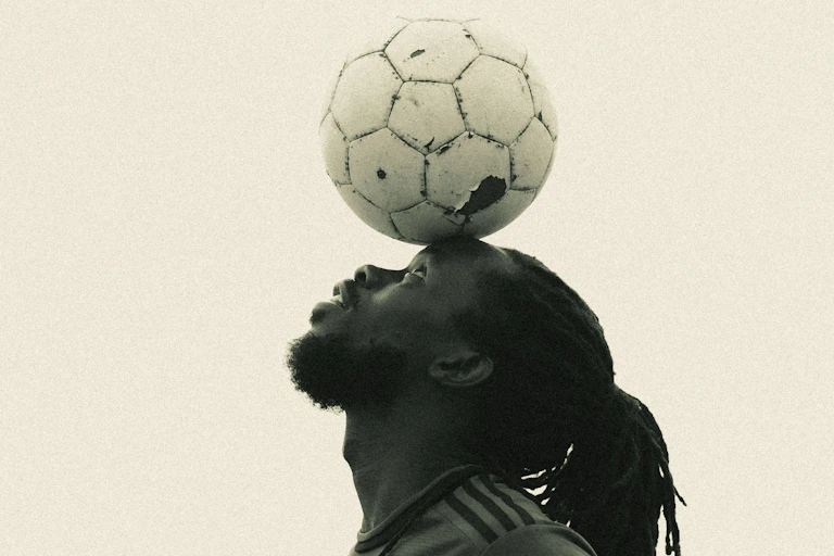 A sepia toned stock photograph of the profile of a young African man with dreadlocks balancing a scuffed football on his forehead.