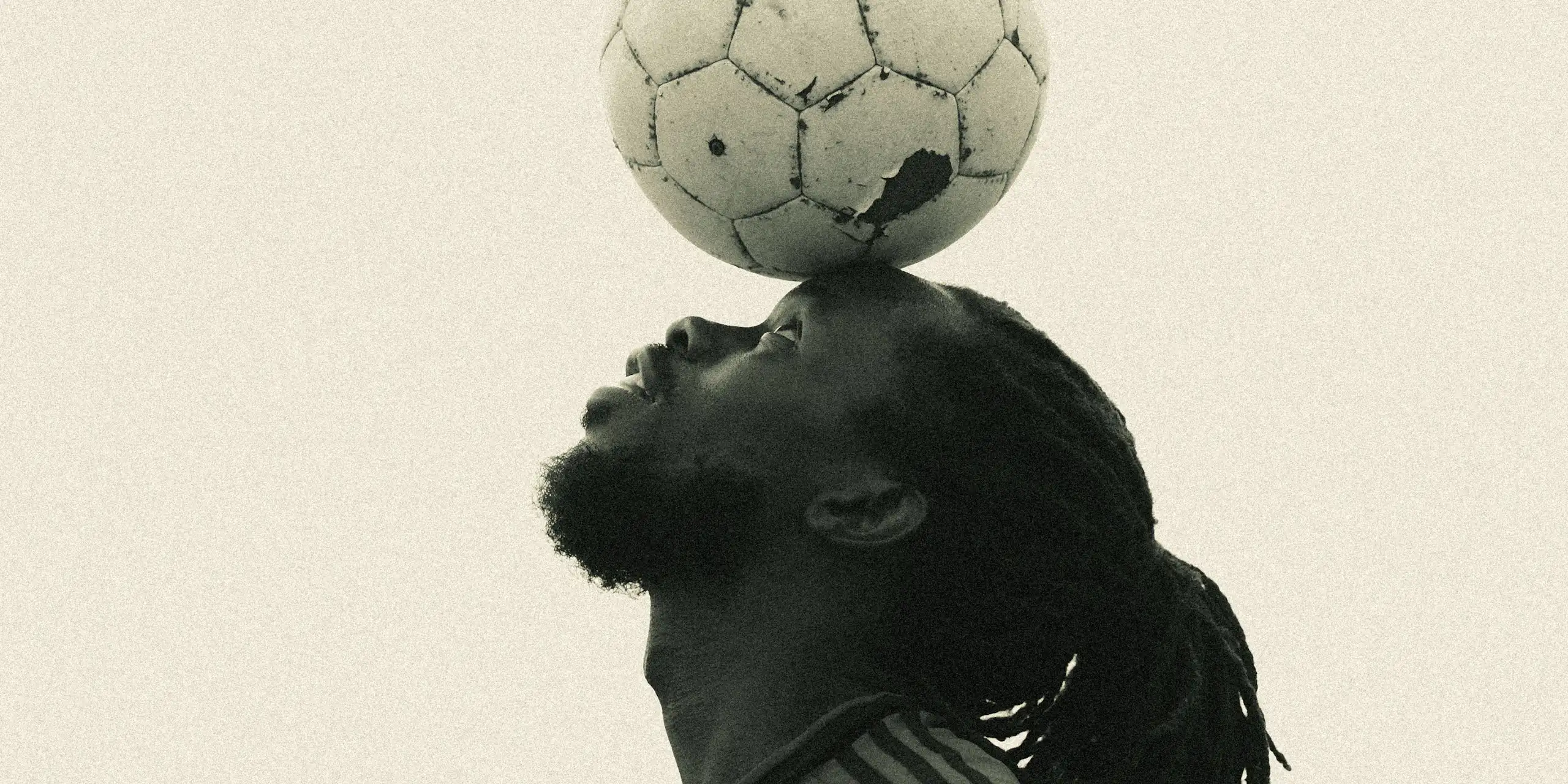 A sepia toned stock photograph of the profile of a young African man with dreadlocks balancing a scuffed football on his forehead.