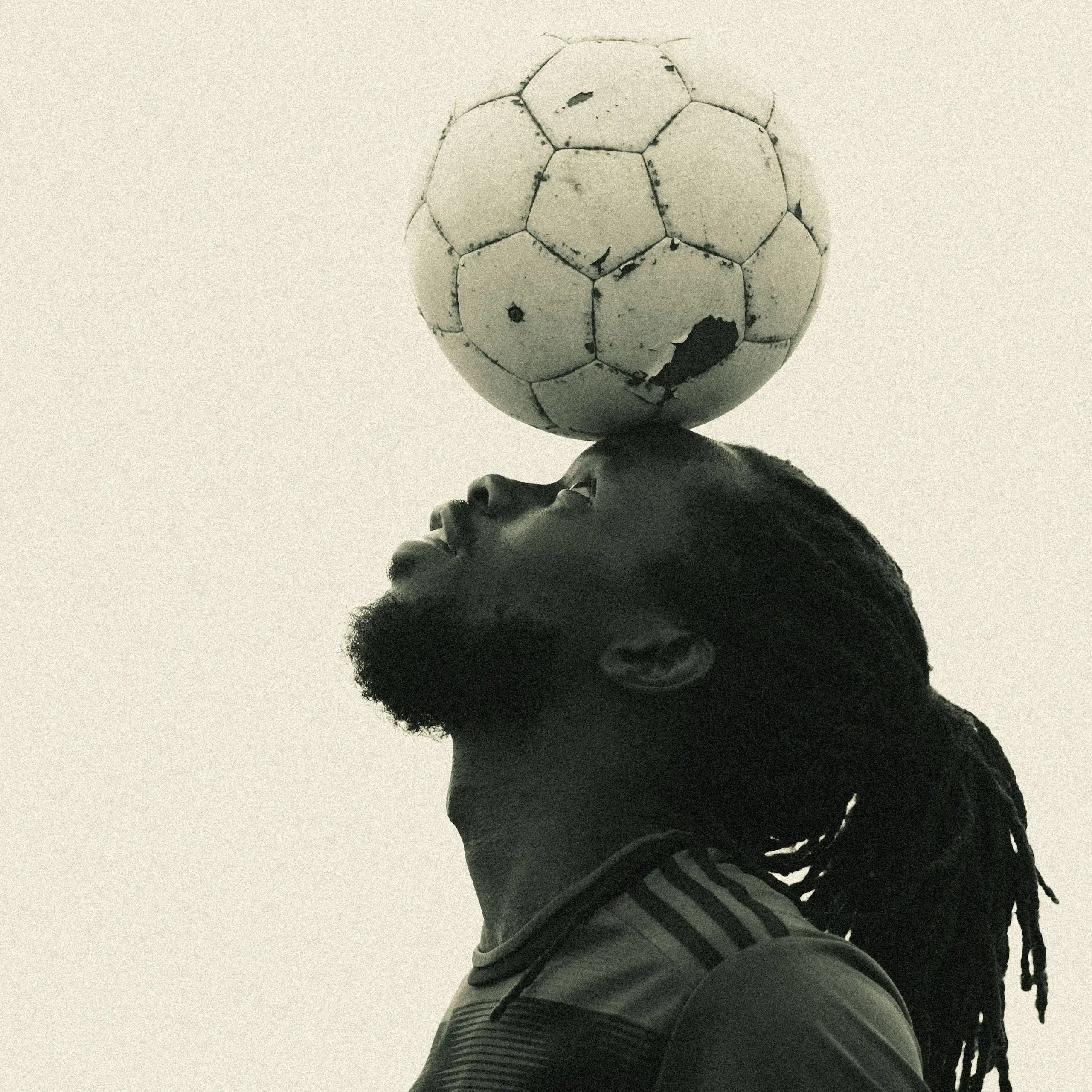 A sepia toned stock photograph of the profile of a young African man with dreadlocks balancing a scuffed football on his forehead.