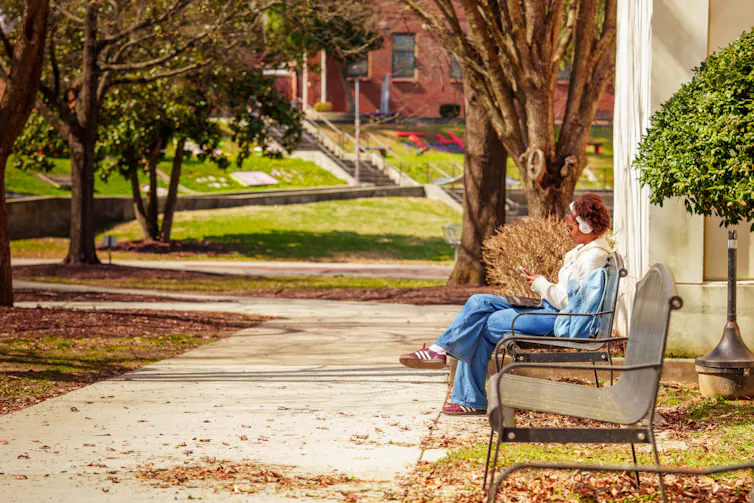 A young Black woman wearing jeans sits on a bench near a sidewalk surrounded by trees and green lawns, with a red building in the distance.