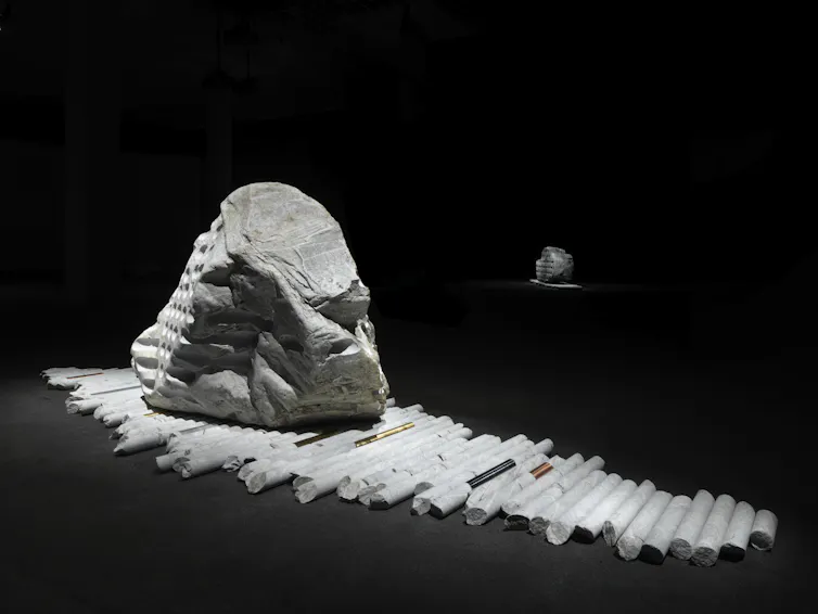 Large glacial erratic boulder displayed on drill cores in a dark gallery. Metal rods placed among the cores reference metal resources extracted from the Earth.