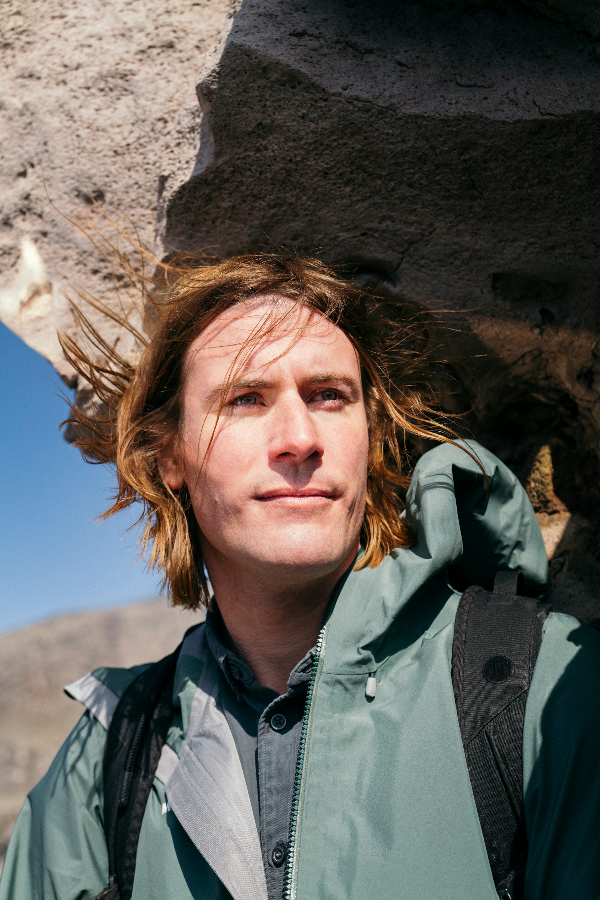 Portrait of artist Julian Charrière standing beside a rocky outcrop in a mountainous landscape. He wears a green jacket and backpack and looks into the distance.