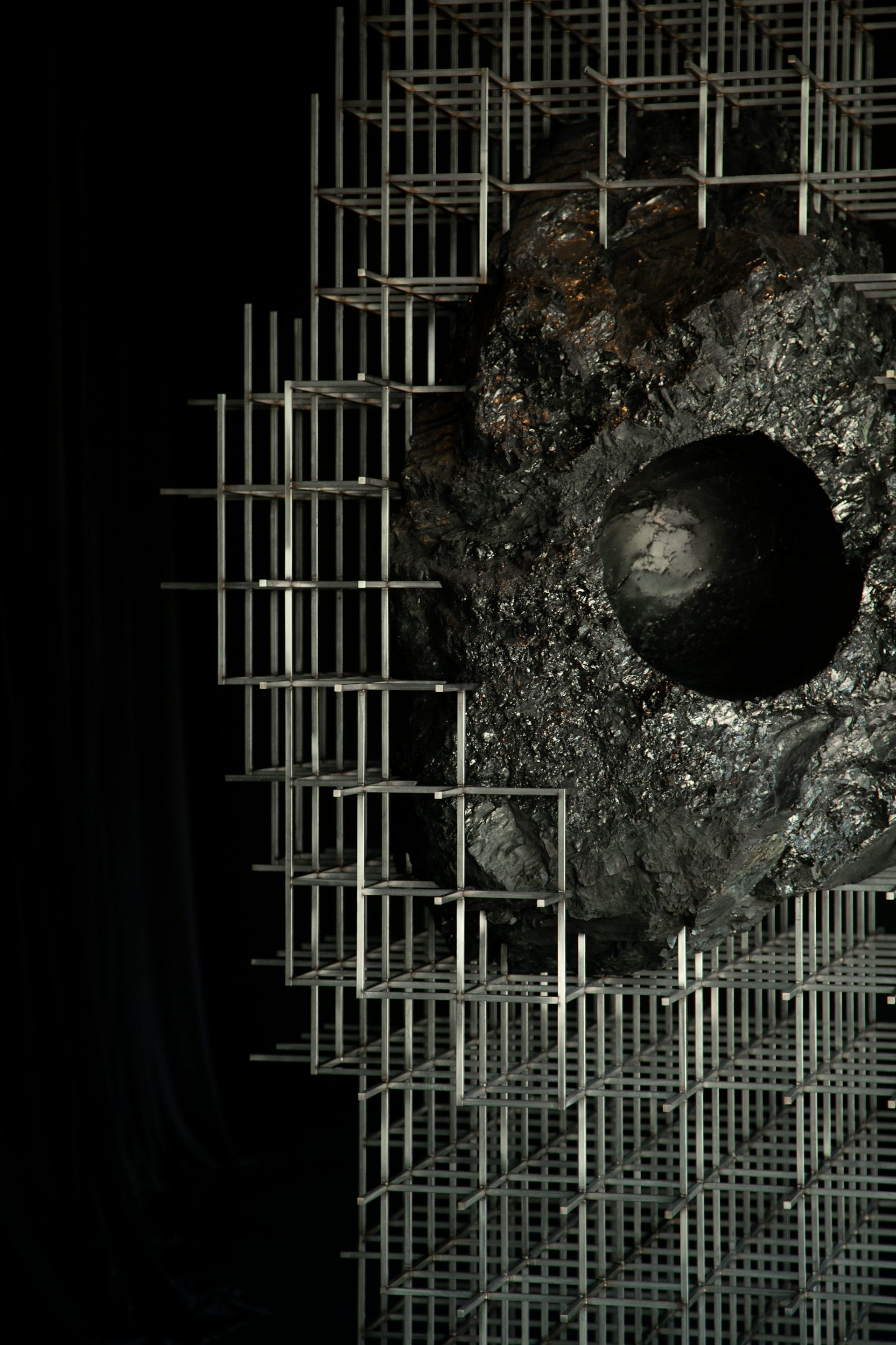 Large black lump of coal suspended within a steel scaffold structure in a dark gallery space. A smooth, head-sized cavity is carved into the coal, inviting visitors to place their head inside the sculpture.