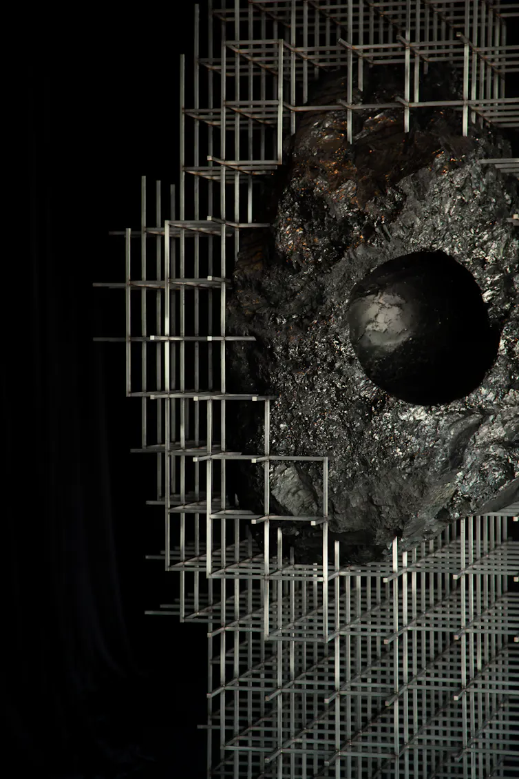 Large black lump of coal suspended within a steel scaffold structure in a dark gallery space. A smooth, head-sized cavity is carved into the coal, inviting visitors to place their head inside the sculpture.