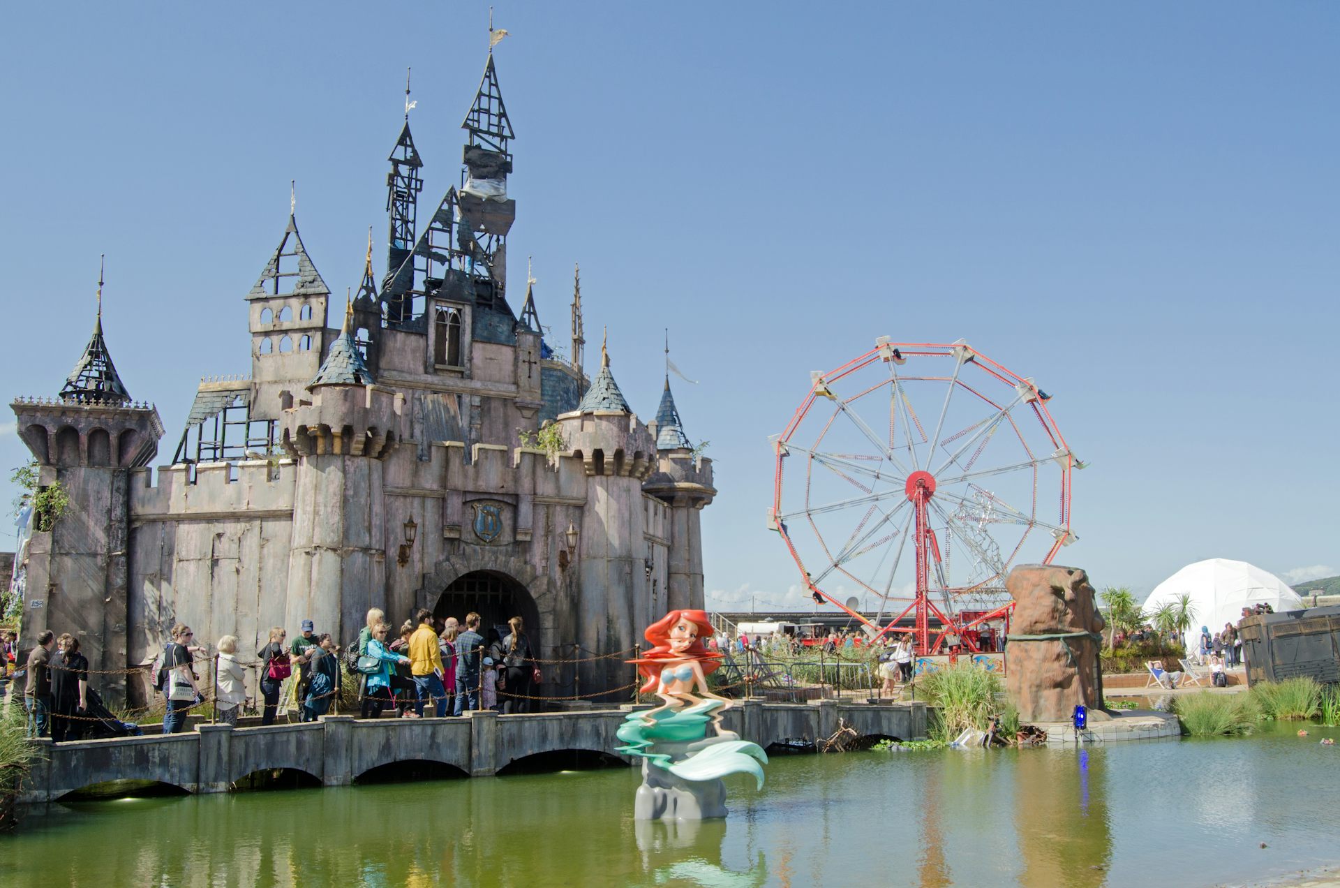 A photograph of the Dismaland theme park, with a distorted Ariel the mermaid sculpture and dark fairytale castle