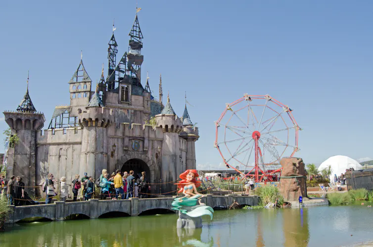 A photograph of the Dismaland theme park, with a distorted Ariel the mermaid sculpture and dark fairytale castle