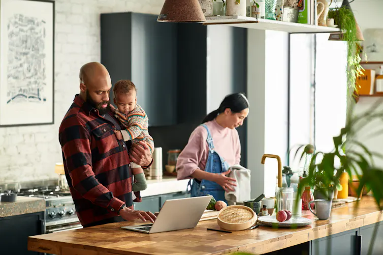 man holding toddler looks at laptop on kitchen island while woman looks down at sink