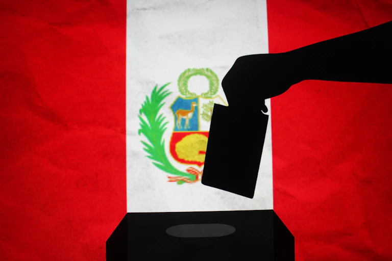 Silueta de una mano introduciendo un voto en una urna con la bandera de Perú al fondo