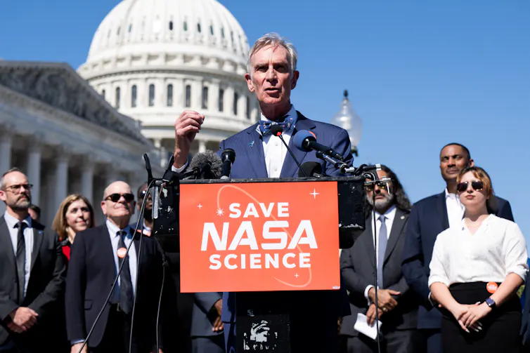 Los recortes a la Fundación Nacional de Ciencias significan que investigadores como yo estamos perdiendo subvenciones, pero los impactos se extienden mucho más allá del mundo académico. 1 Un hombre mayor blanco con una chaqueta azul y pajarita se encuentra en un podio con el cartel
