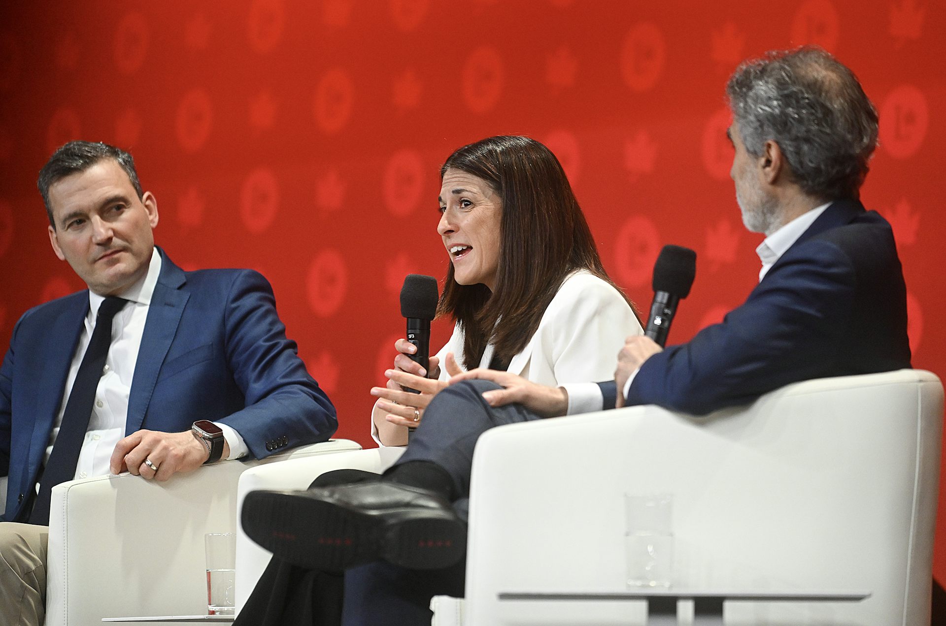 Two men and a women sit in white armchairs while speaking on a stage