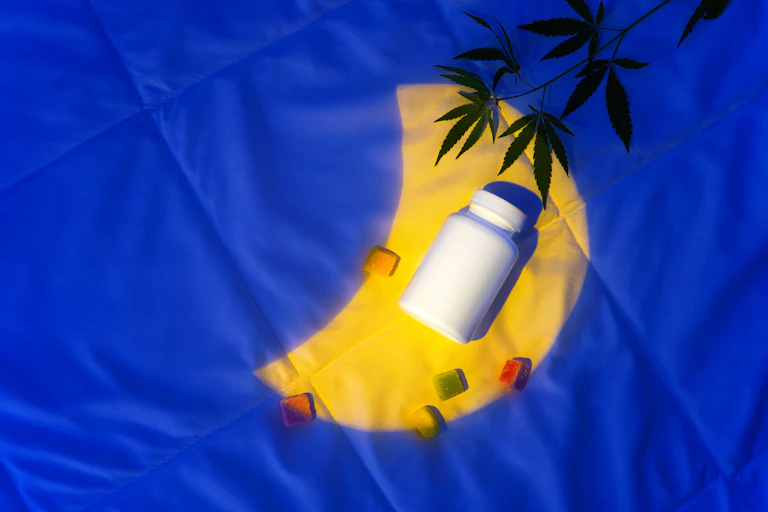 Projected image of a half-moon on a bedspread, with a cannabis plant, a handful of gummies and a pill bottle lying on it.