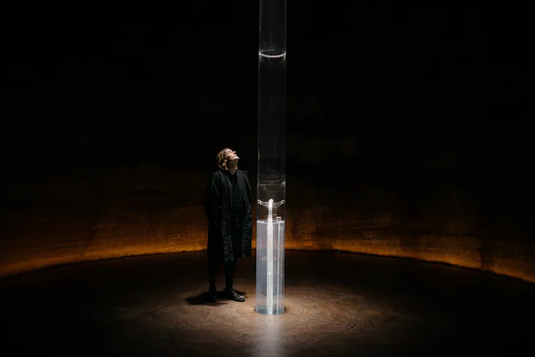 Artist Julian Charrière stands beside a tall transparent column inside a circular, dimly lit chamber. The illuminated installation, Breathe, extends from floor to ceiling and is designed to release oxygen extracted from ancient rocks.