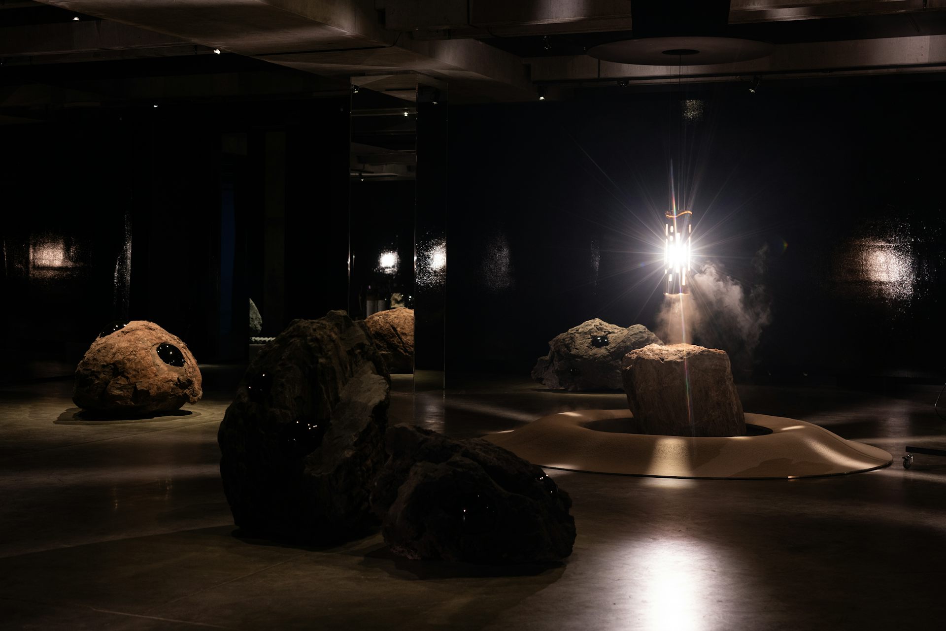 Installation view of Hard Core by Julian Charrière at MONA. Large volcanic rocks and lava bombs are arranged in a dark, mirrored gallery around a bright central light source, creating the impression of a volcanic chamber.