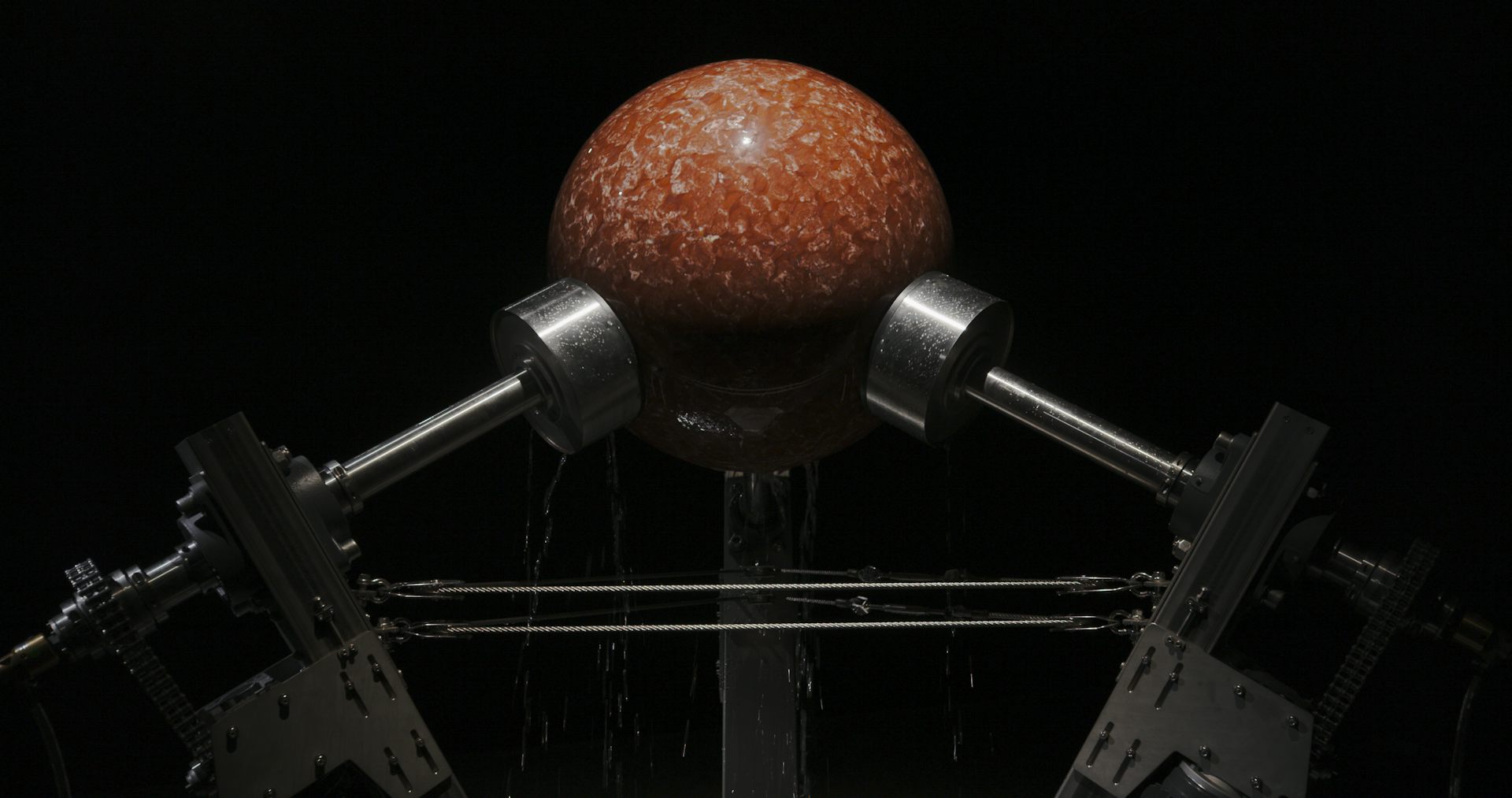 Ancient stromatolite fossil held between metal rollers in a mechanical framework. Water flows over the rock as the machine gradually grinds it into a smooth spherical shape.