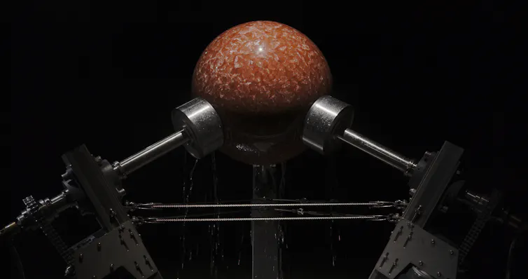 Ancient stromatolite fossil held between metal rollers in a mechanical framework. Water flows over the rock as the machine gradually grinds it into a smooth spherical shape.
