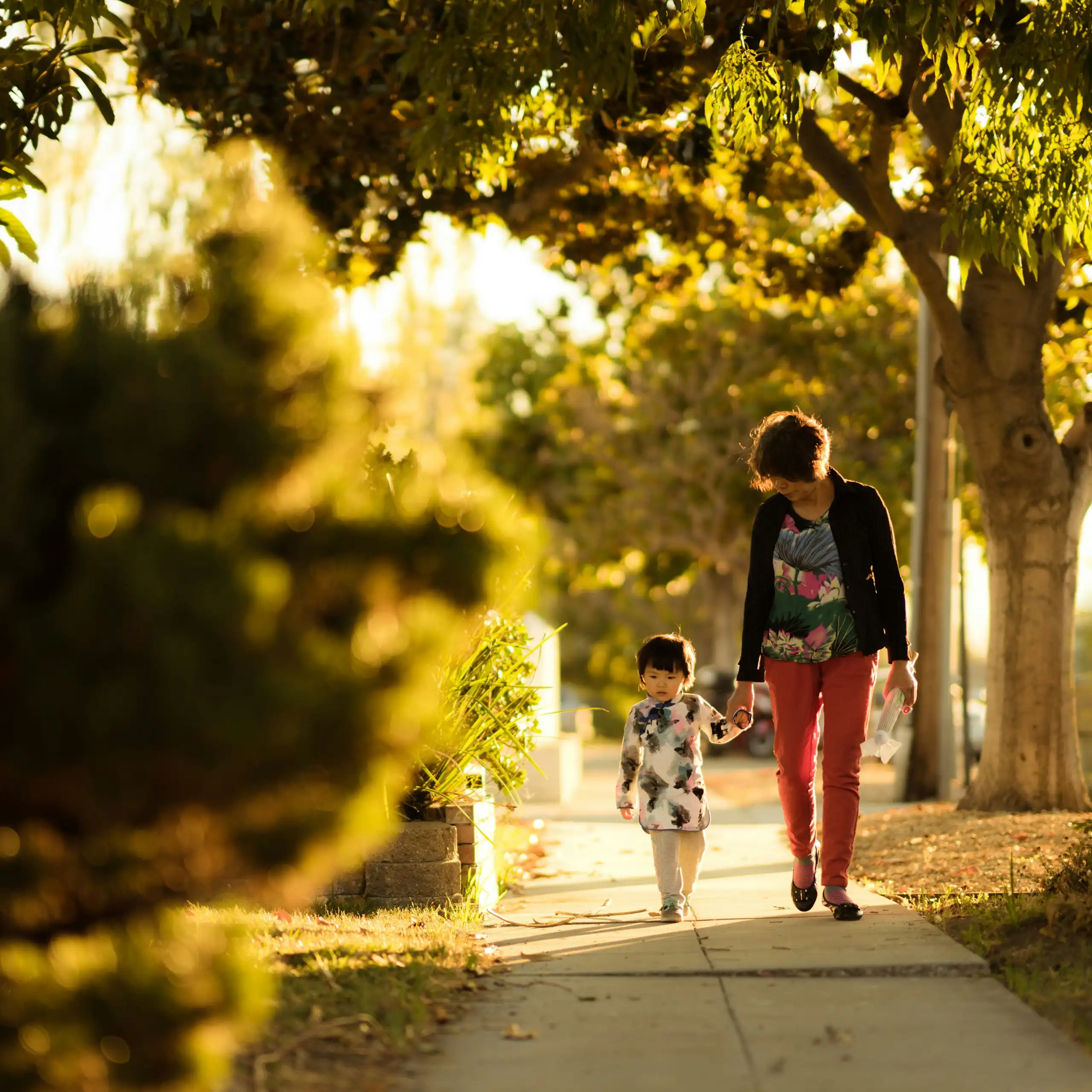 A mother holds her young daughter's hand as they walk down the path in the sun