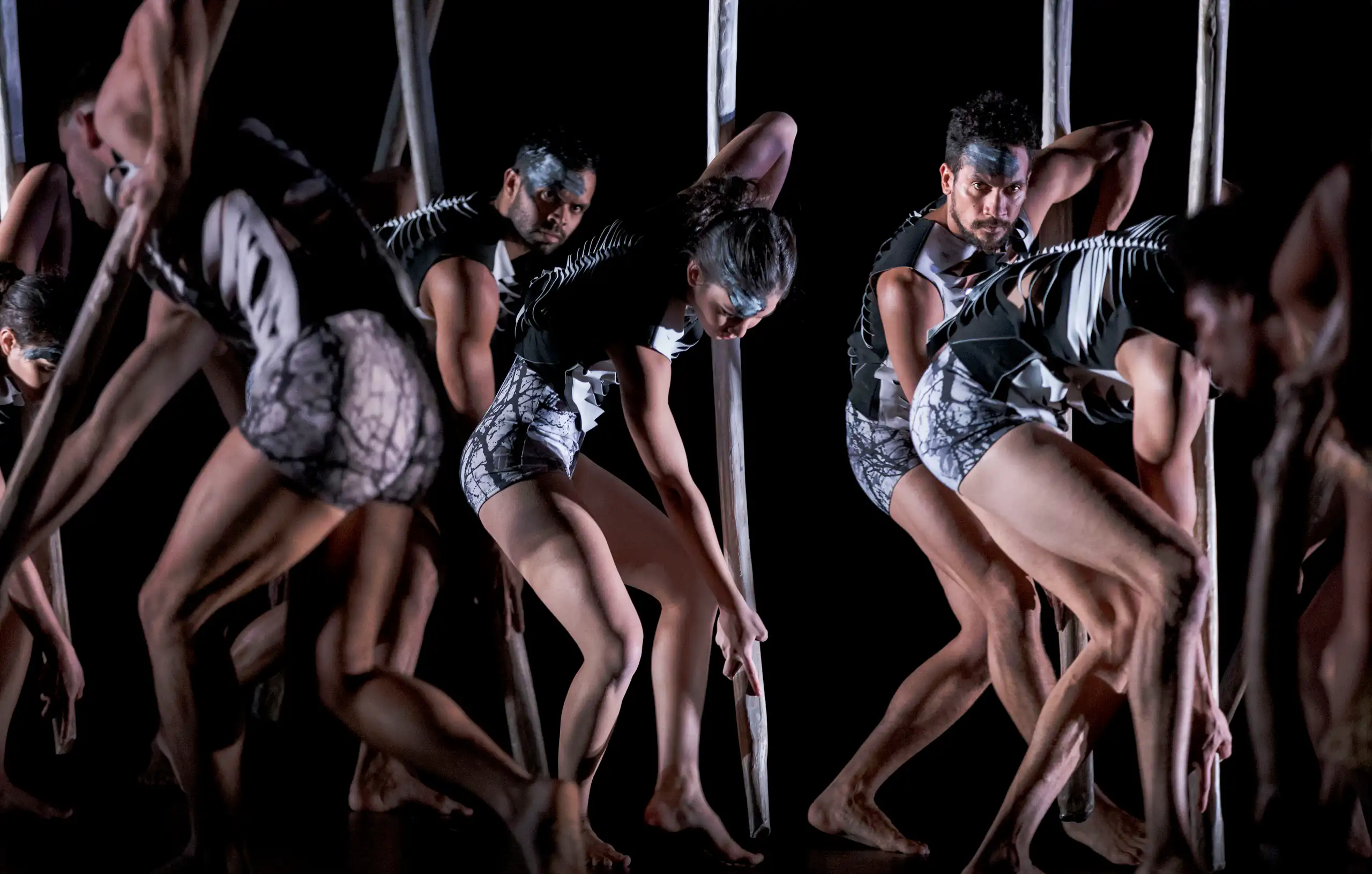 An ensemble of dancers in skeleton-like outfits march across a dark stage.