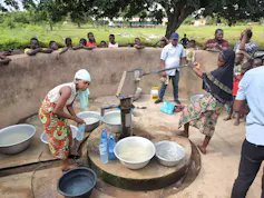 A group of people stands around a borehole collecting water in basins