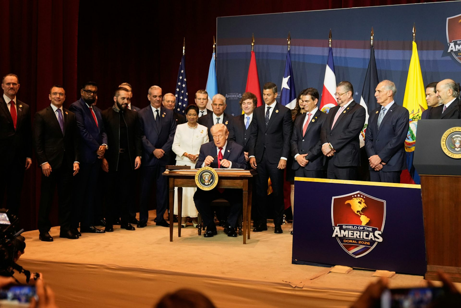 A gathering of world leaders in suits stand around a man at a desk.