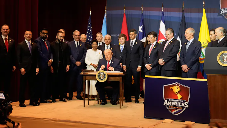 A gathering of world leaders in suits stand around a man at a desk.