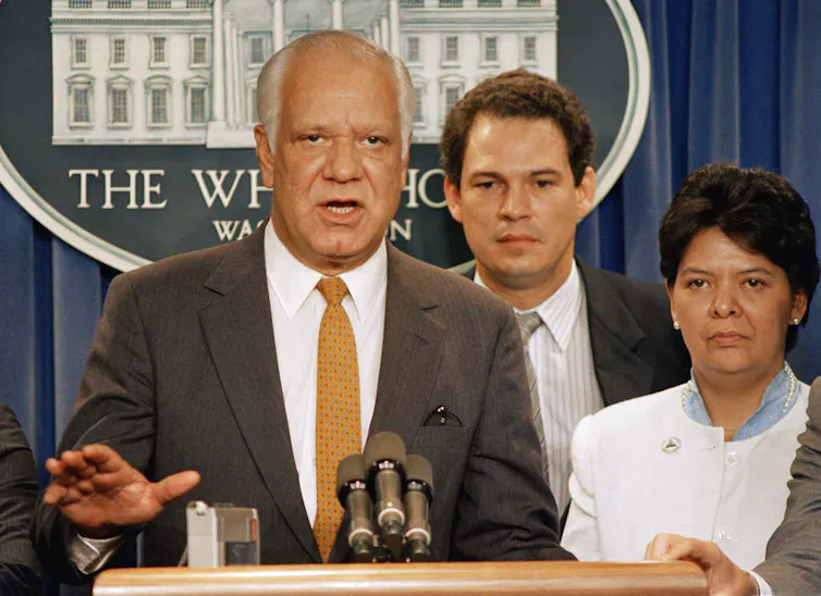 A man stands at a lectern alongside two others.