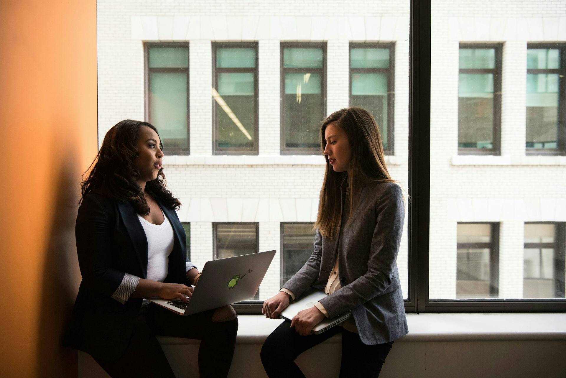 Two women are talking, sitting on a windowsill, each with a laptop on her lap