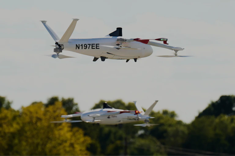 two small windowless aircraft in the air with a tree line in the background