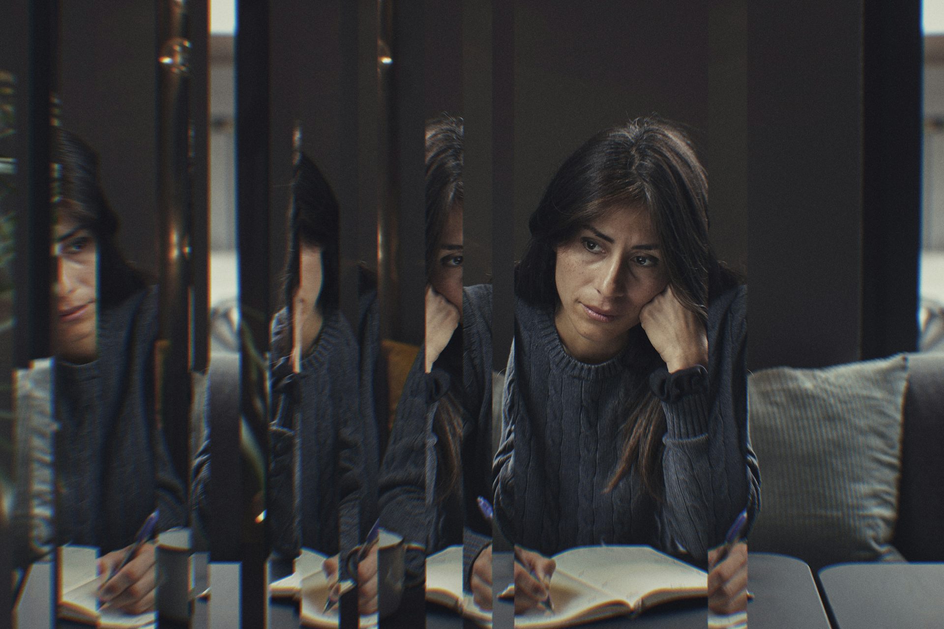 Una morena escribe en un diario y parece pensativa, su rostro parcialmente reflejado en varios finos paneles de vidrio.