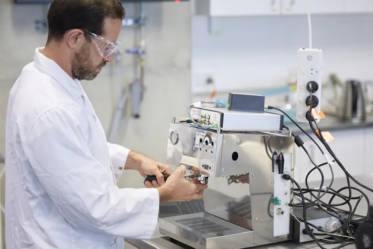 Man in white lab coat and goggles stands at an espresso machine with some kind of electronic box attached.