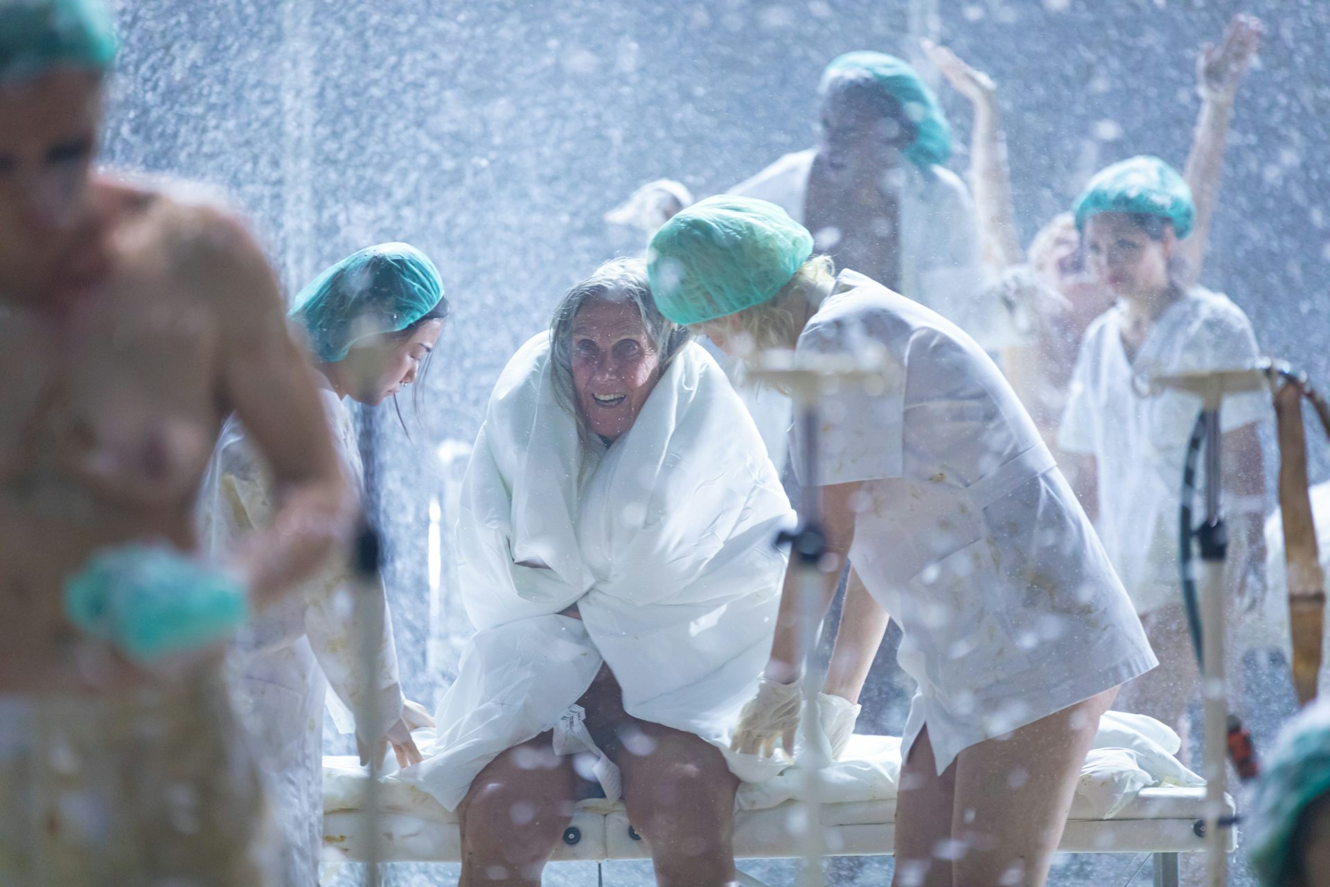 An older performer is onstage in a hospital setting. She has a blanket wrapped around her and seems to be shivering.