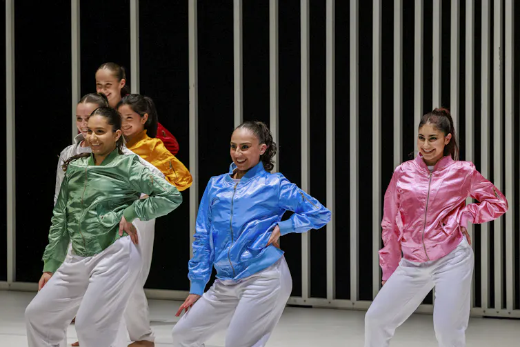 Women dancers in brighly coloured jackets pose with their hands on their hips mid-dance.