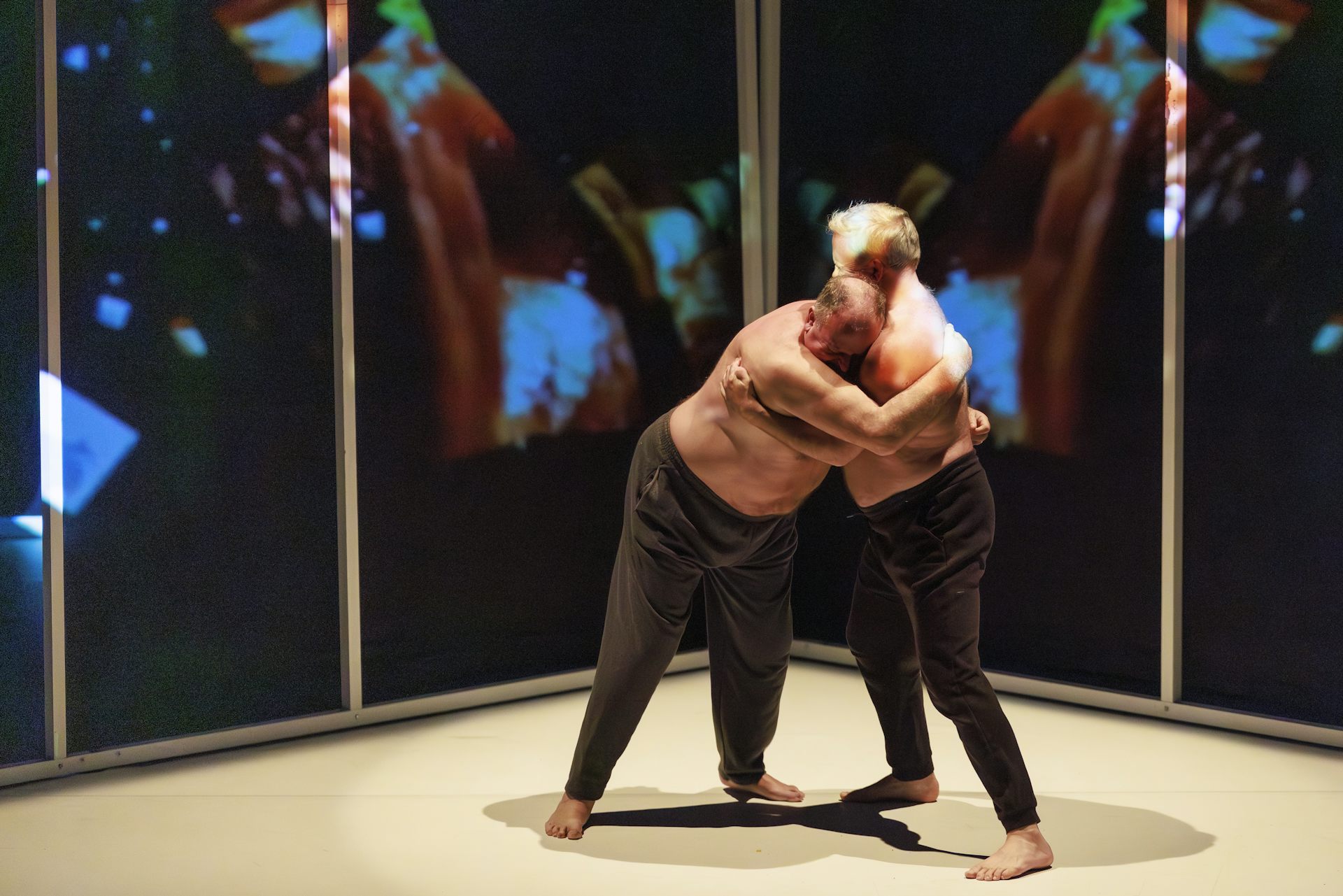 Two shirtless performers wrap their arms around each other during a dance performance.