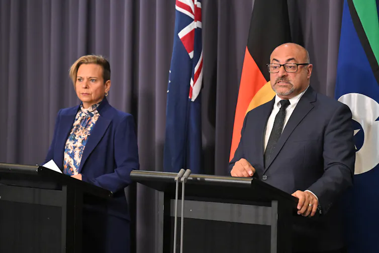 two people standing at podiums in front of Australian flags.