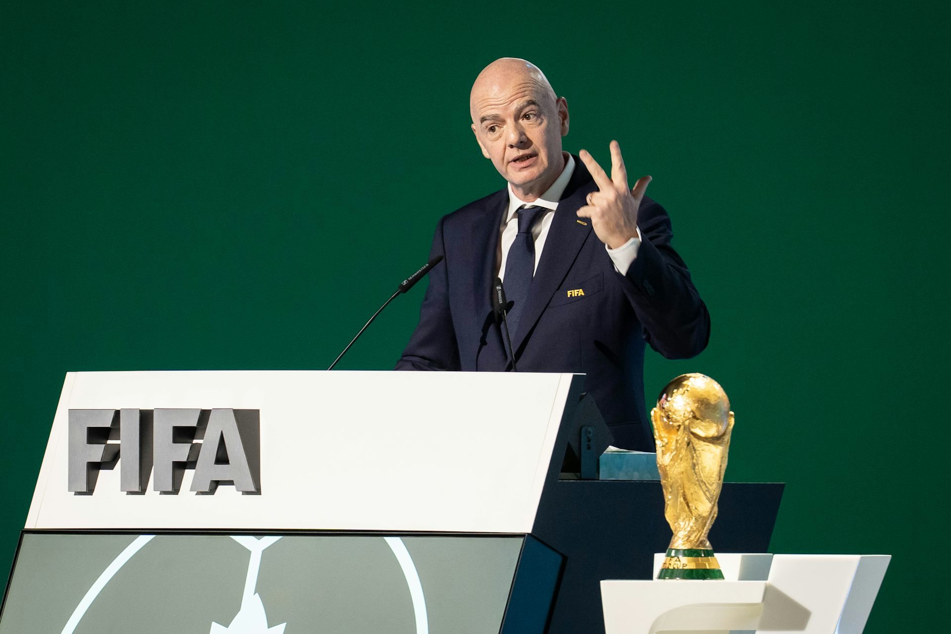 un hombre blanco calvo con traje habla en un podio con un cartel de la FIFA y un trofeo dorado a su lado