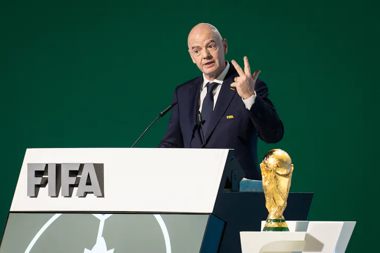 un hombre blanco calvo con traje habla en un podio con un cartel de la FIFA y un trofeo dorado a su lado