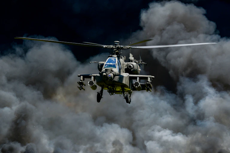 A US Army Apache helicopter against the backdrop of a smoke-filled sky.