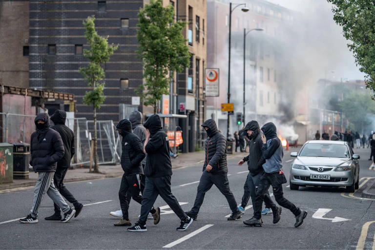hooded youths on the streets of belfast with smoke rising in the background from a burning shop.
