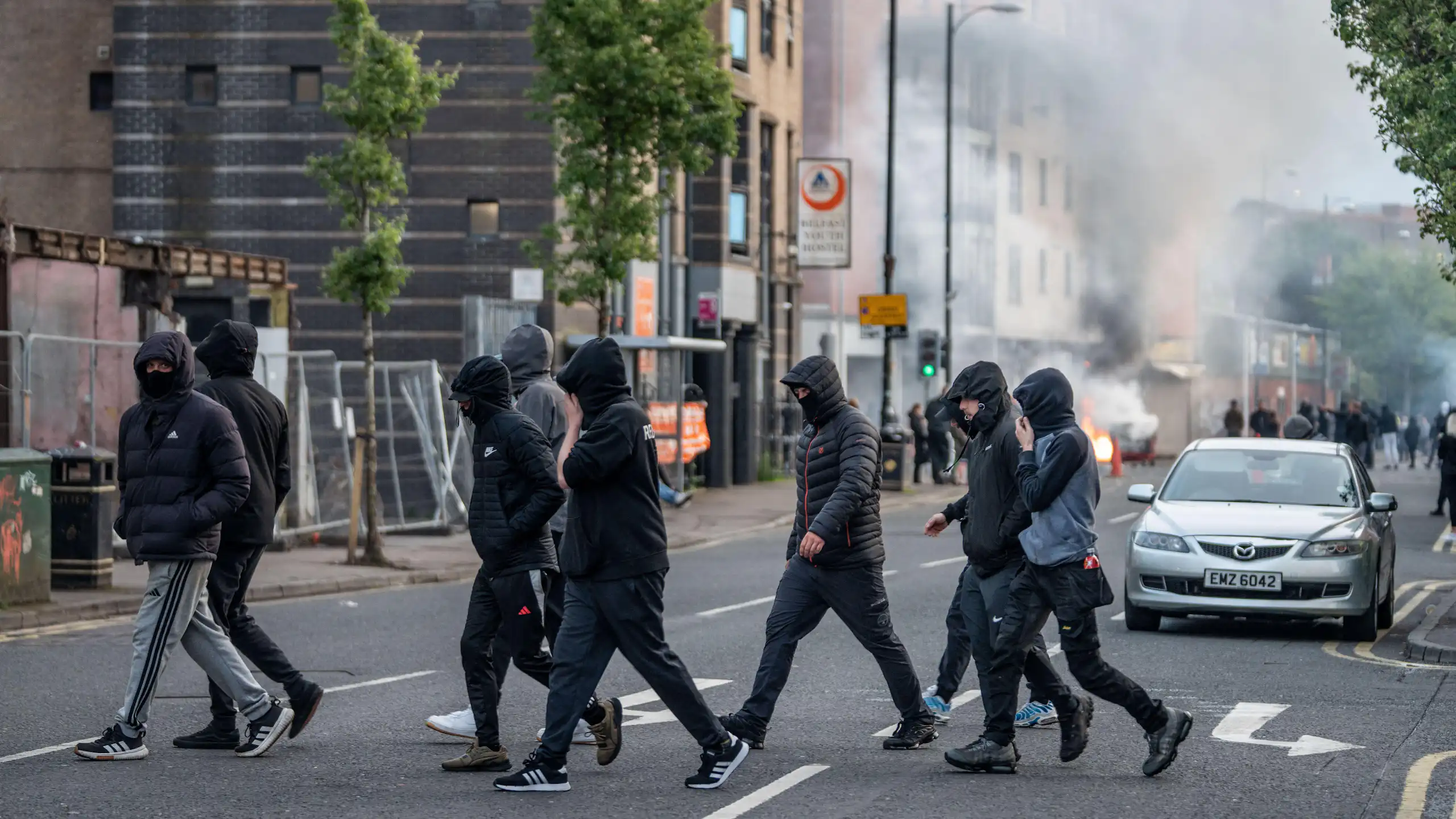 hooded youths on the streets of belfast with smoke rising in the background from a burning shop.