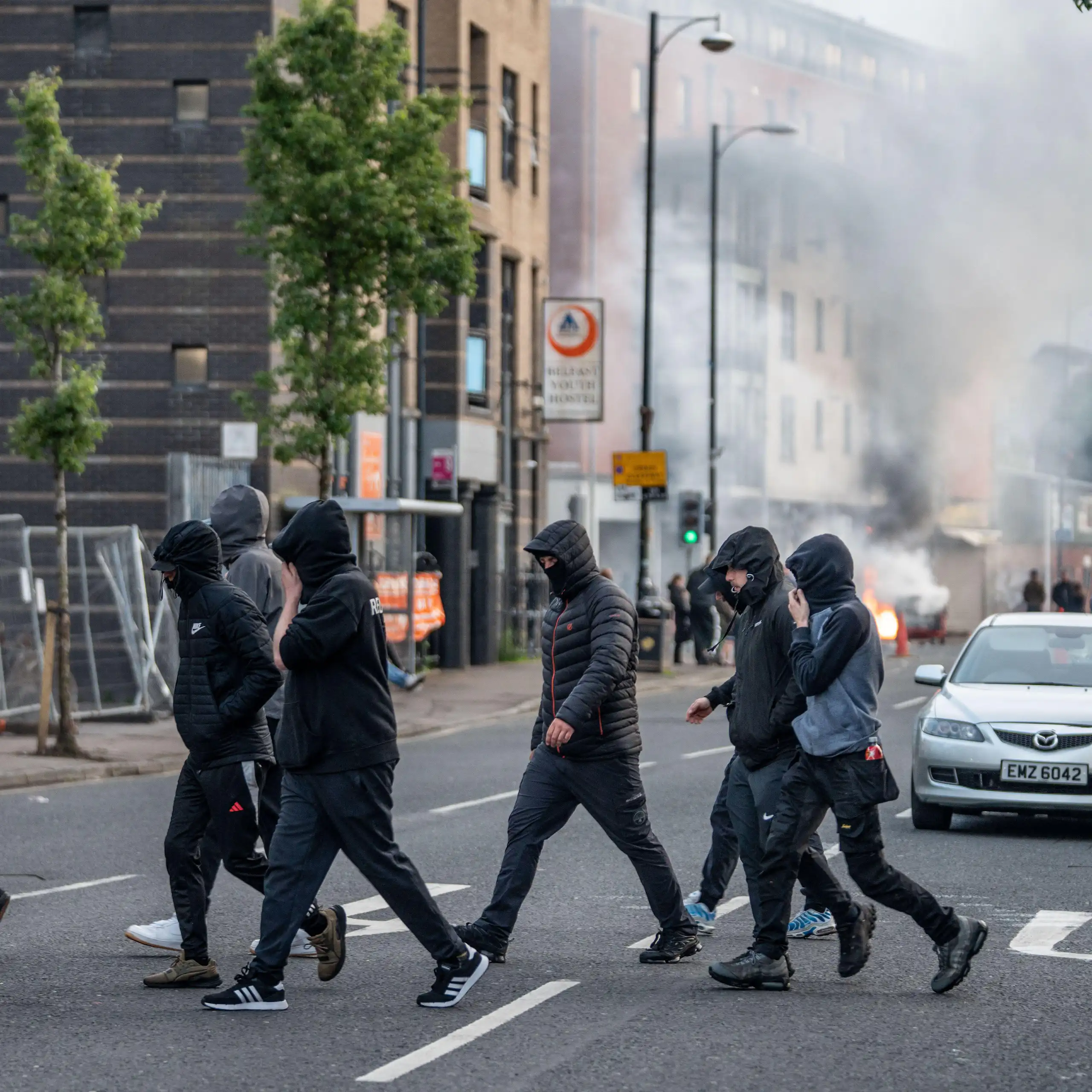 hooded youths on the streets of belfast with smoke rising in the background from a burning shop.
