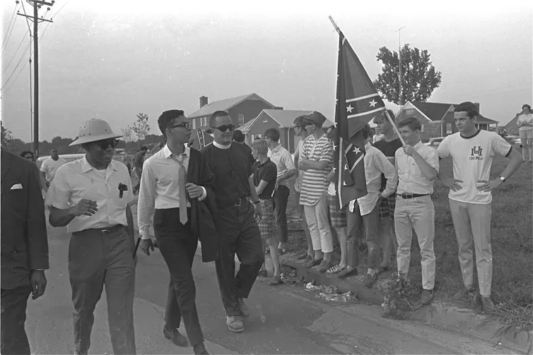 A Black man walks by white men, one of them holding a Confederate flag.
