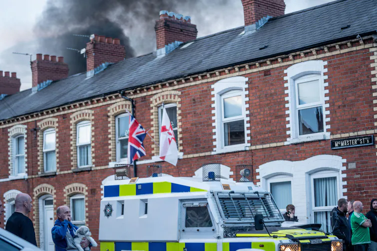 row of terraced houses with police riot van in front of them and smoke rising behind them