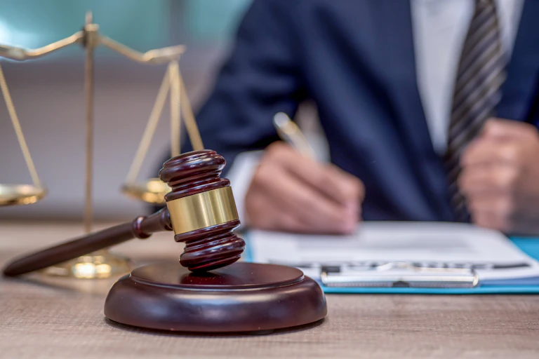 A gavel and scale sit in front of a person in a blue suit with a pen in their hand.