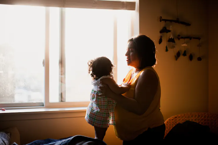 A mom and holds her toddler child as they stand in a room in front of a sun-filled window.