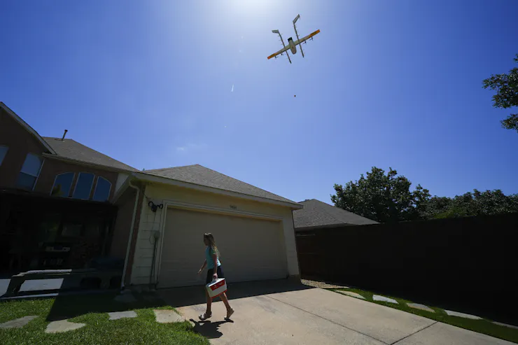 a girl carries a package in a house's driveway as a small aircraft flies above
