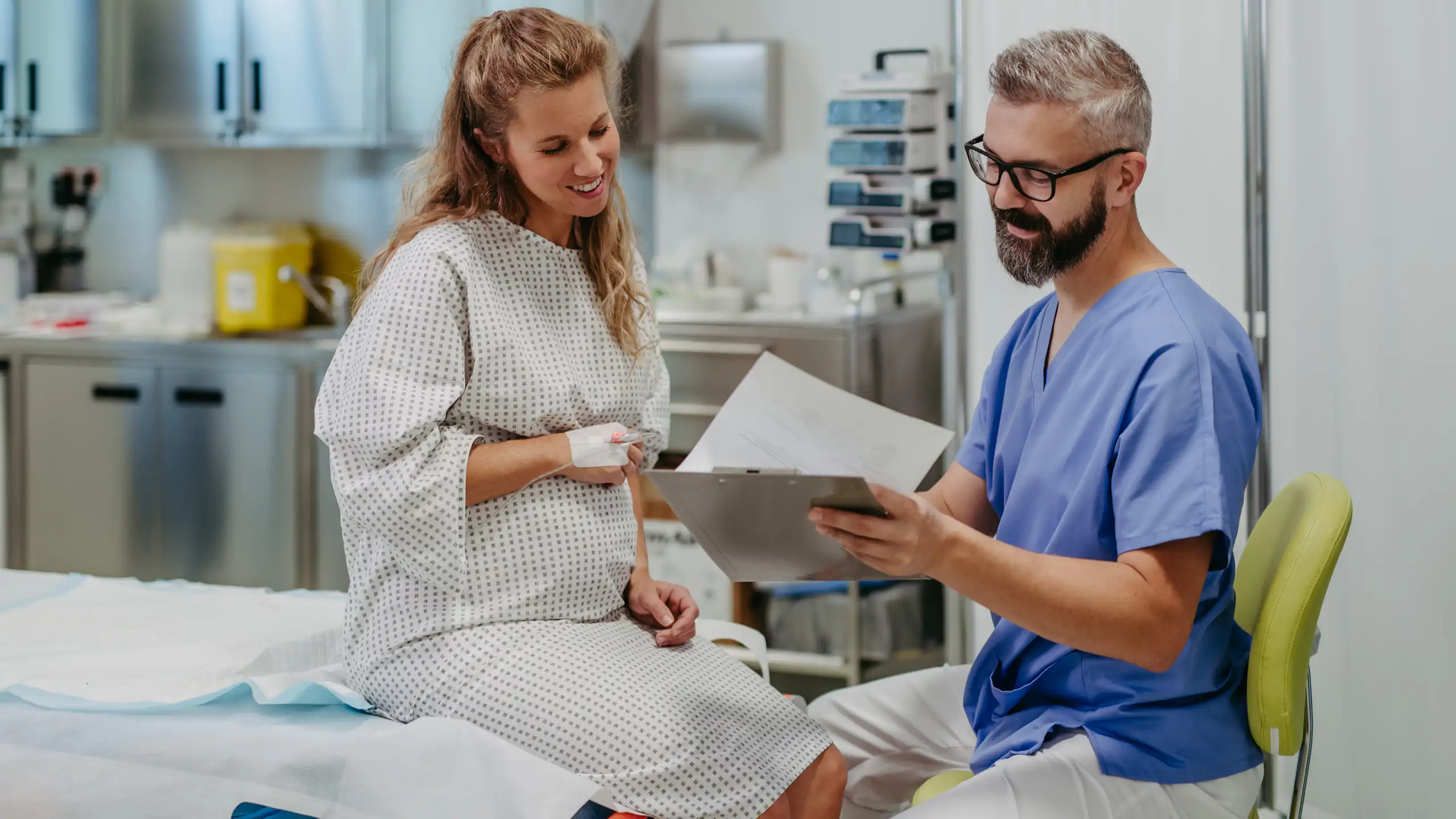 A pregnant woman sits in a hospital gown and chats with a man in scrubs.