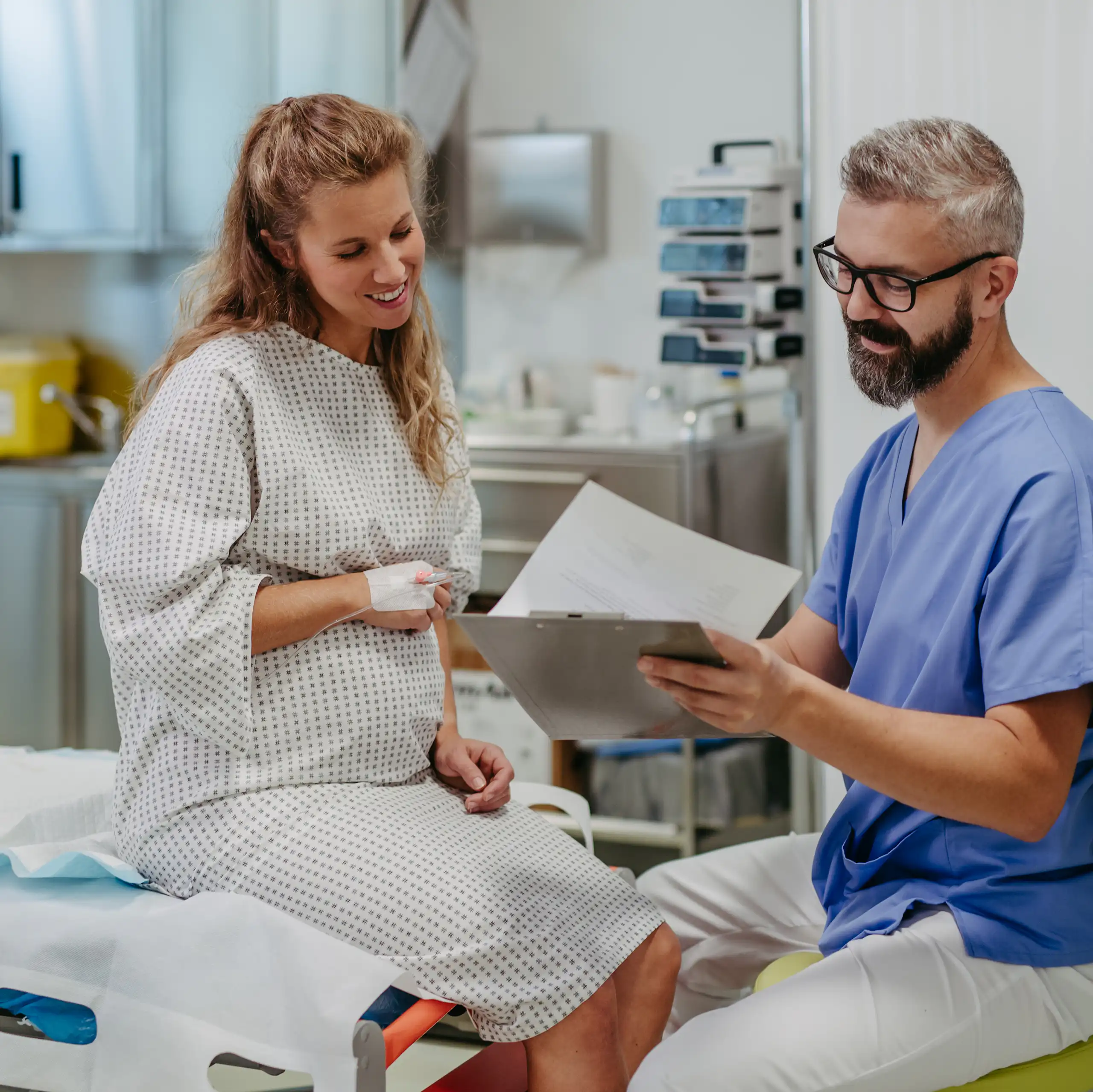 A pregnant woman sits in a hospital gown and chats with a man in scrubs.